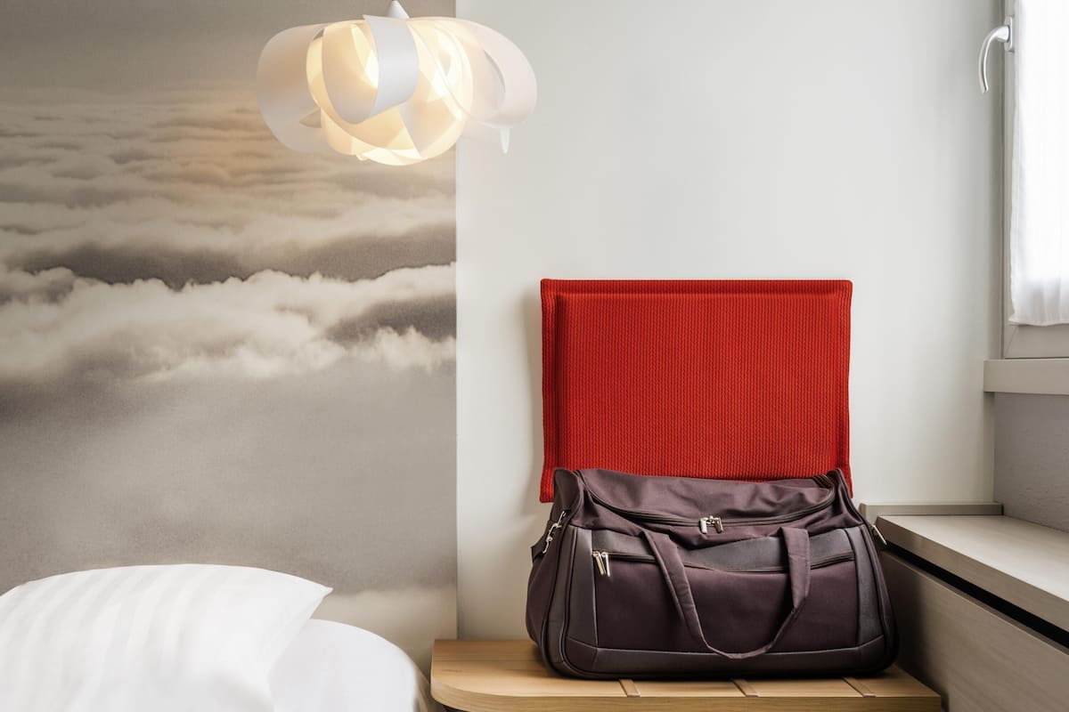 Una habitación con una cama, un panel de pared rojo, una lámpara colgante y un banco de madera con un bolso encima.