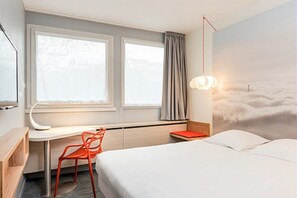 Una habitación de hotel con cama, escritorio, silla, ventana con cortinas y un mural en la pared.