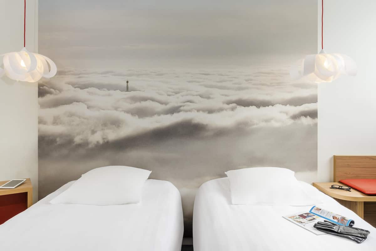 Habitación de hotel con dos camas, un gran mural con motivos de nubes en la pared y una mesita de noche de madera con una tableta y gafas.