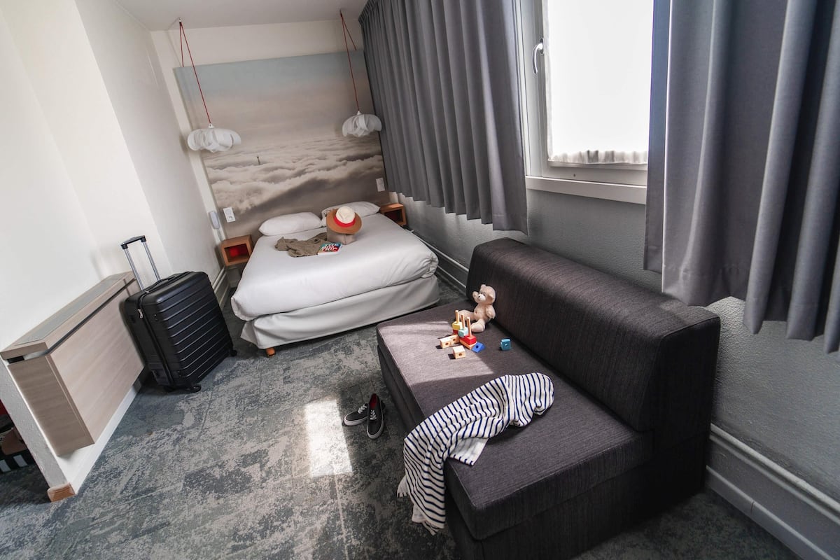 Una habitación de hotel con cama, sofá, maleta, oso de peluche y ventana con cortinas.
