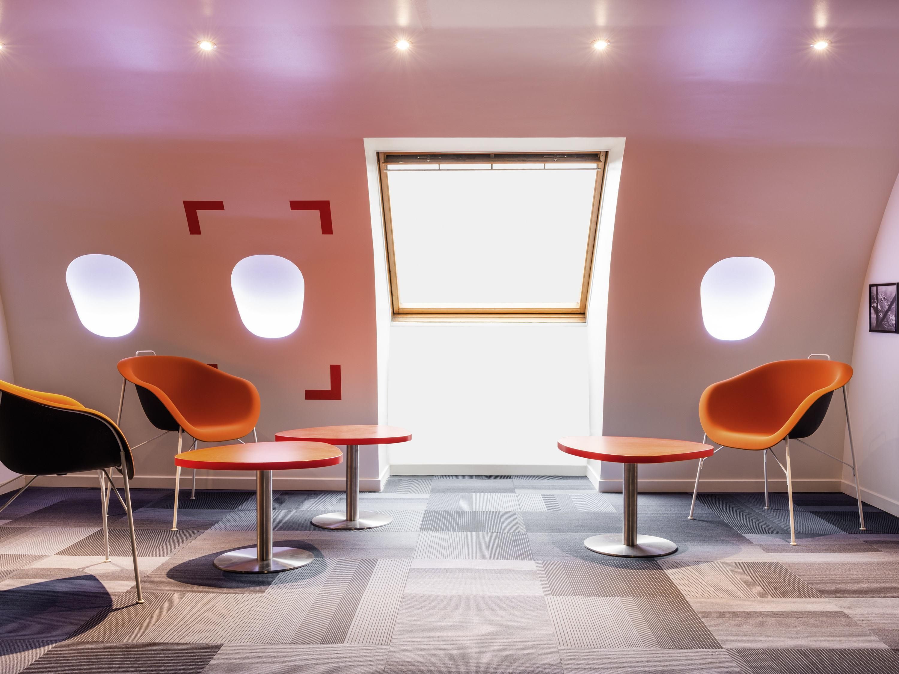 Un área de espera moderna con dos sillones naranjas, una mesa de madera y un ventanal grande.