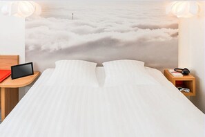 A bed with a view of clouds, a bedside table with a tablet, and a nightstand with headphones. - ibis Styles Paris Velizy (Velizy-Villacoublay)