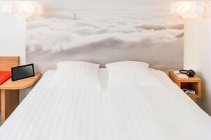 Una cama con vistas a las nubes, una mesita de noche con una tableta y un mueble de noche con auriculares.