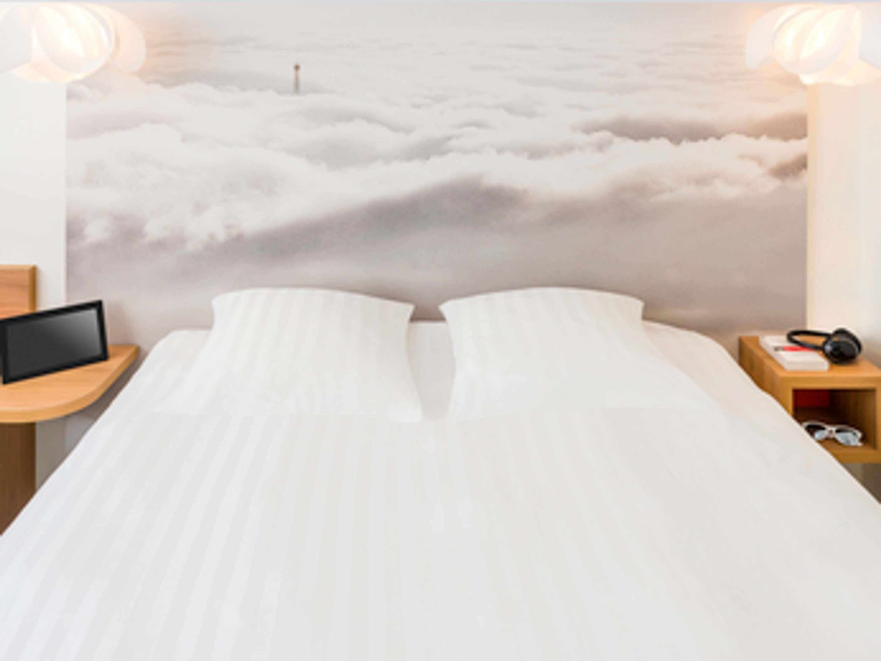 A bed with a white mattress and pillows, flanked by wooden nightstands with a tablet and headphones.