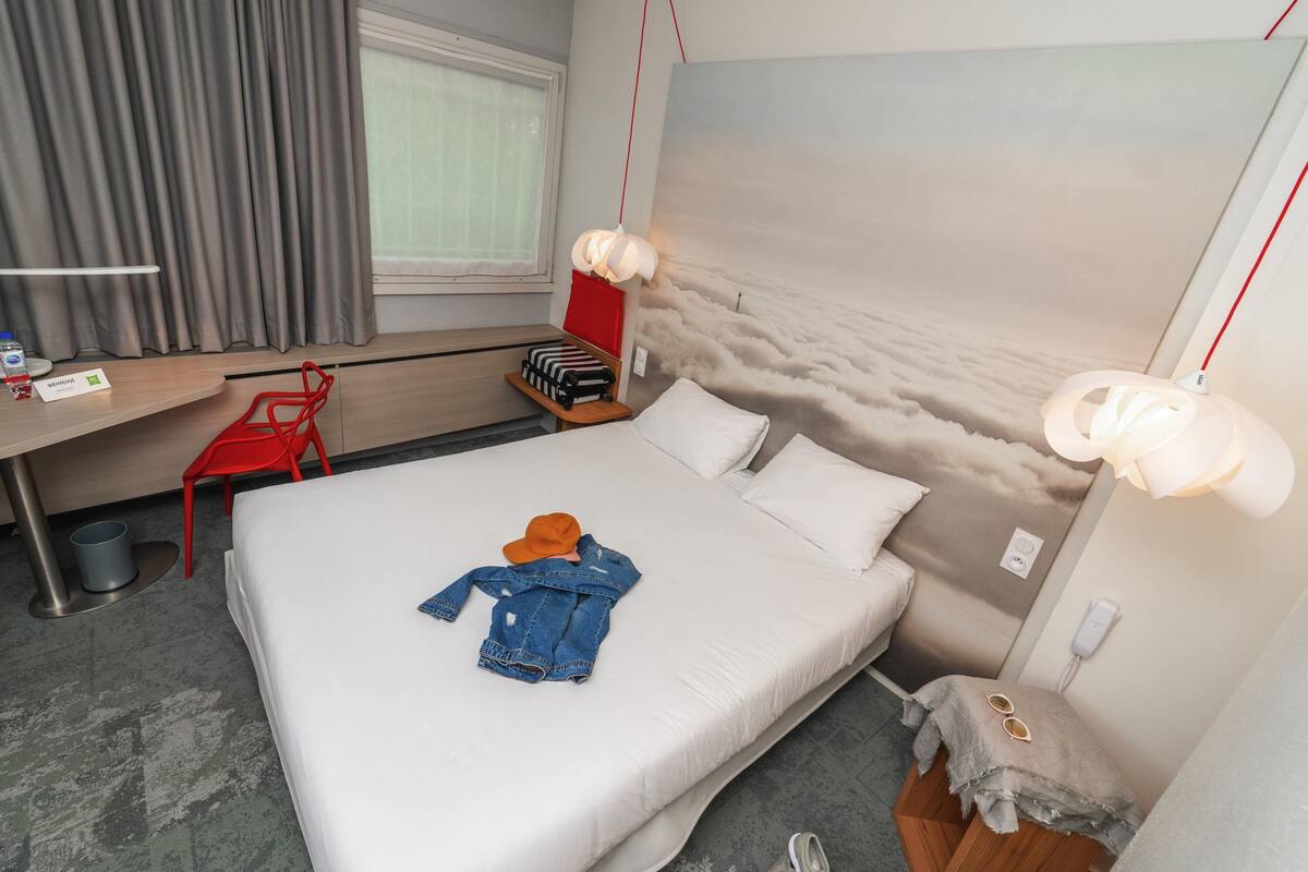 Una habitación de hotel con cama, escritorio, silla, maleta y un mural en la pared.