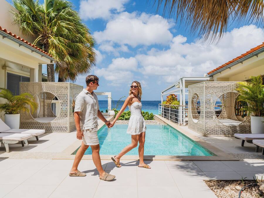 Un couple se promène près d’une piscine, où se trouvent des chaises longues et des palmiers.