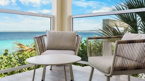A balcony with wicker chairs and a small table, overlooking a turquoise sea and greenery.