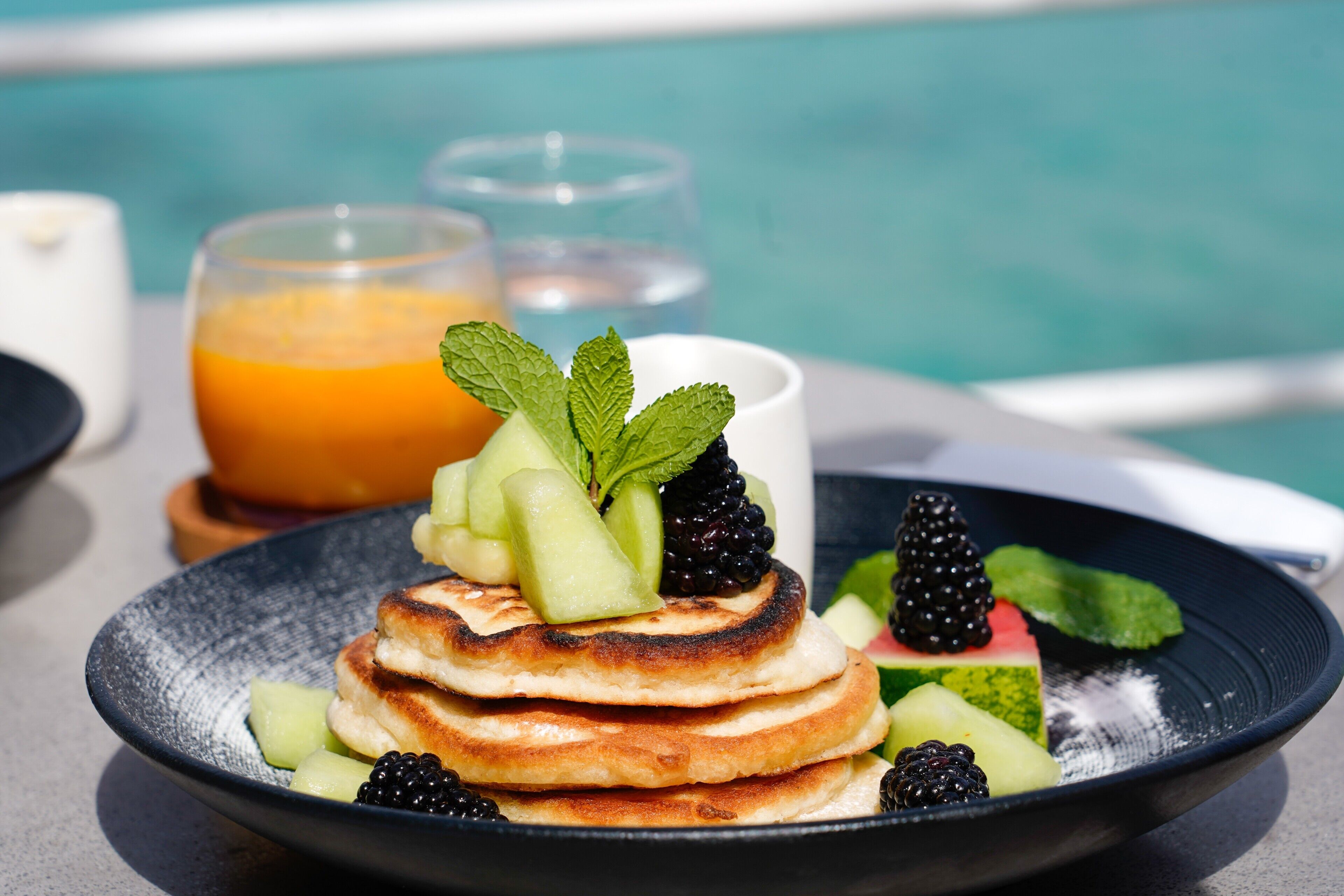 Un plato de desayuno con panqueques, moras y melón, acompañado de un vaso de jugo de naranja y una taza blanca, con vista a una piscina.