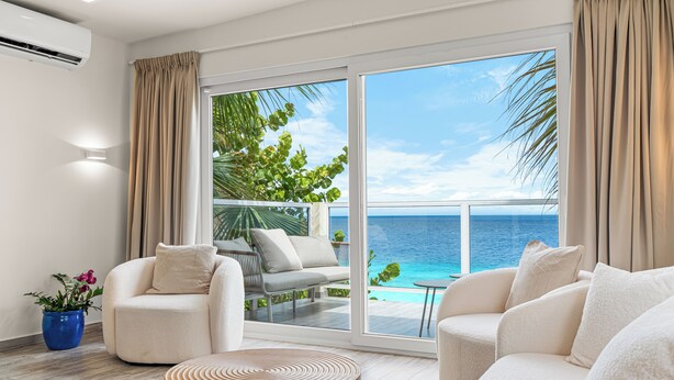 A modern living room with a large sliding glass door leading to a balcony with a view of the ocean. There are two white sofas, a round wooden coffee table, and a blue potted plant.