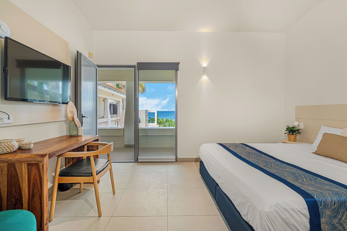 A hotel room with a bed, a desk, a chair, a TV, and a sliding door leading to a balcony with a view of the sea.