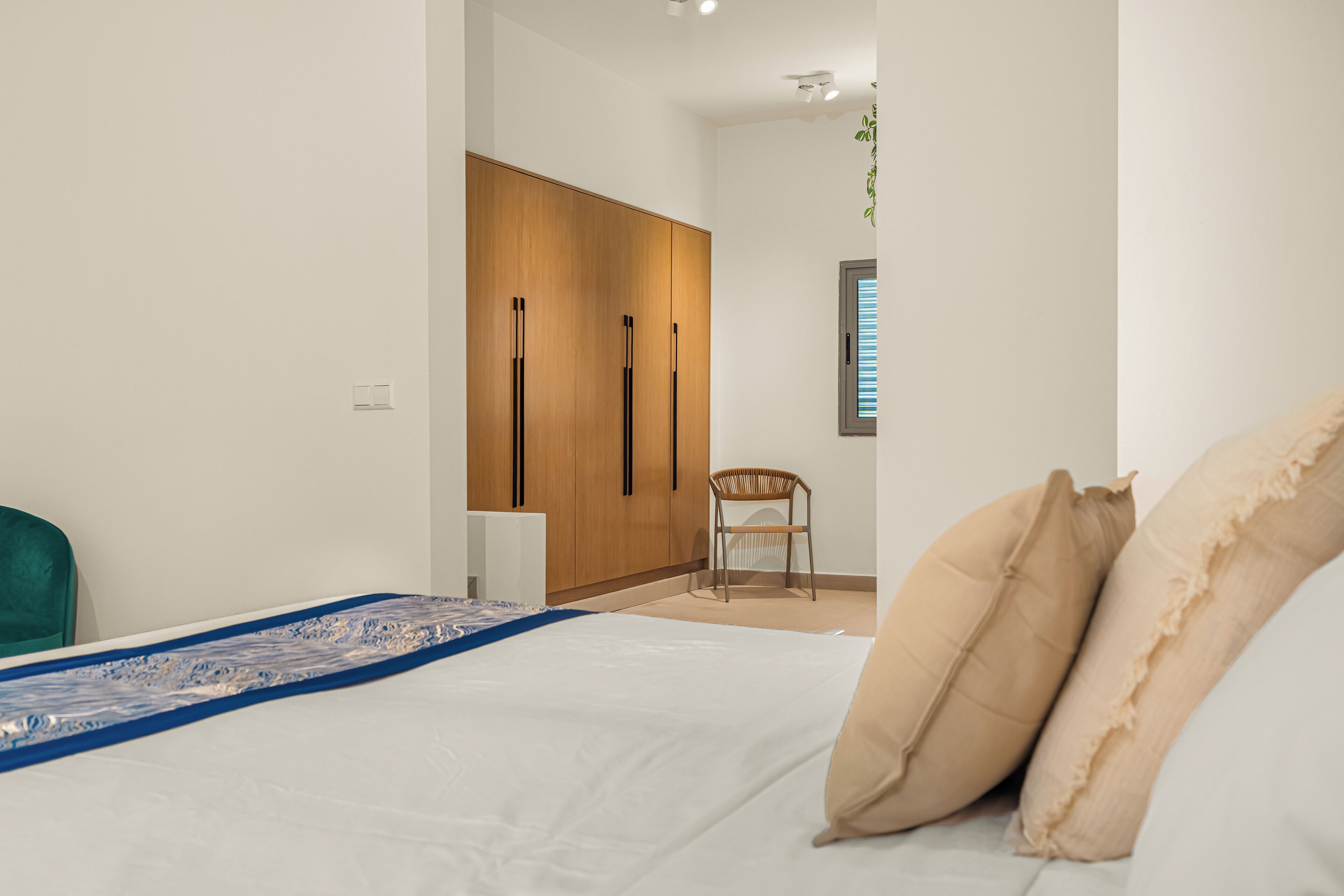 A bedroom with a bed, a wooden wardrobe, a chair, and a window with blinds.