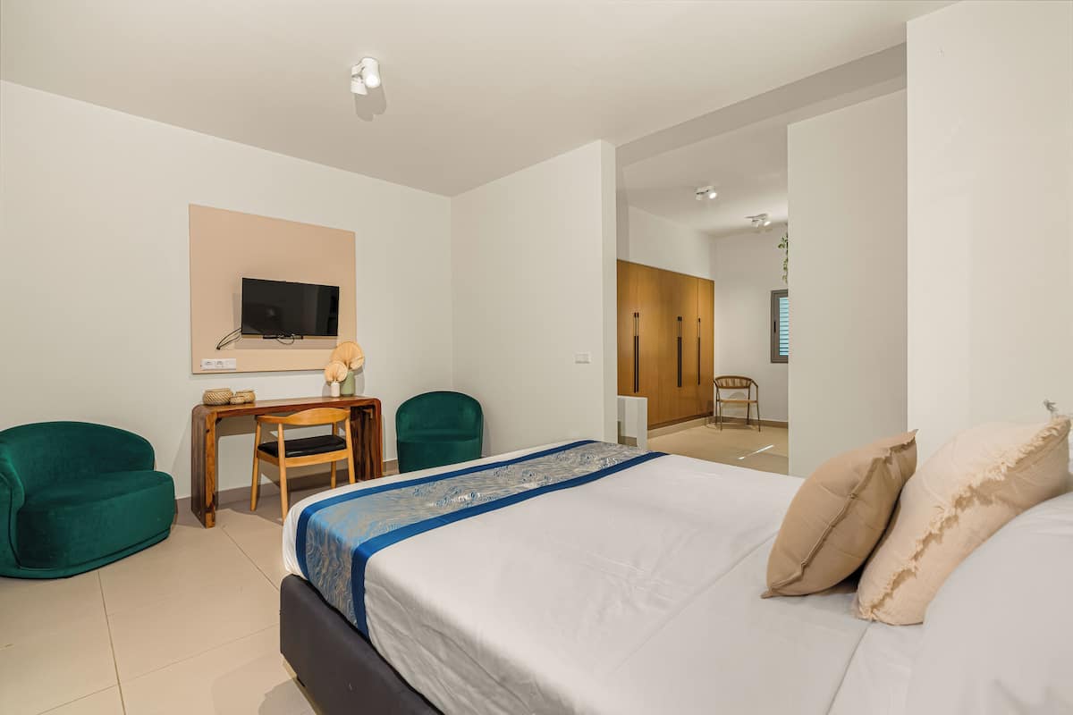 A modern hotel room with a bed, two green chairs, a small wooden table, and a television on the wall.