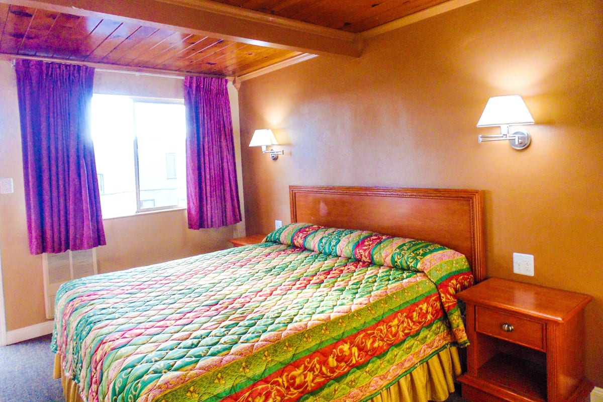 A bedroom with a wooden ceiling, a bed with a colorful quilt, purple curtains, and two wall-mounted lamps.