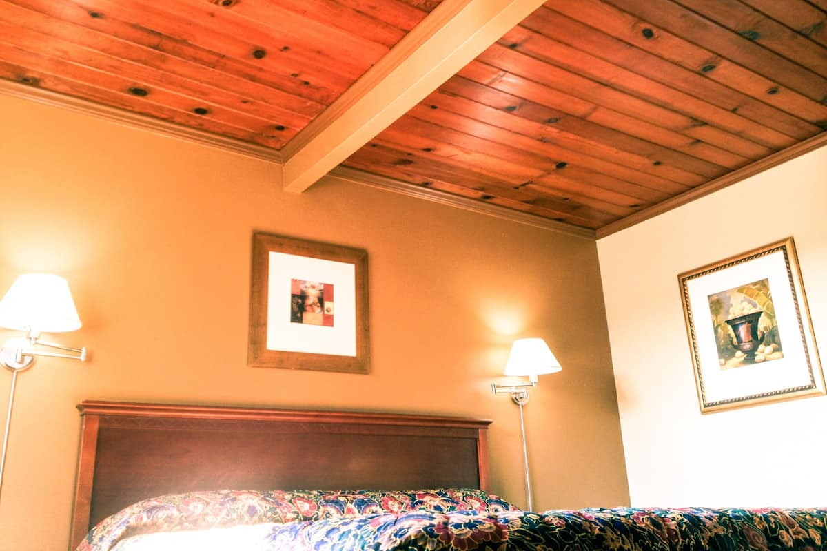 A room with a wooden ceiling, a bed with a floral bedspread, two wall-mounted lamps, and two framed pictures on the walls.