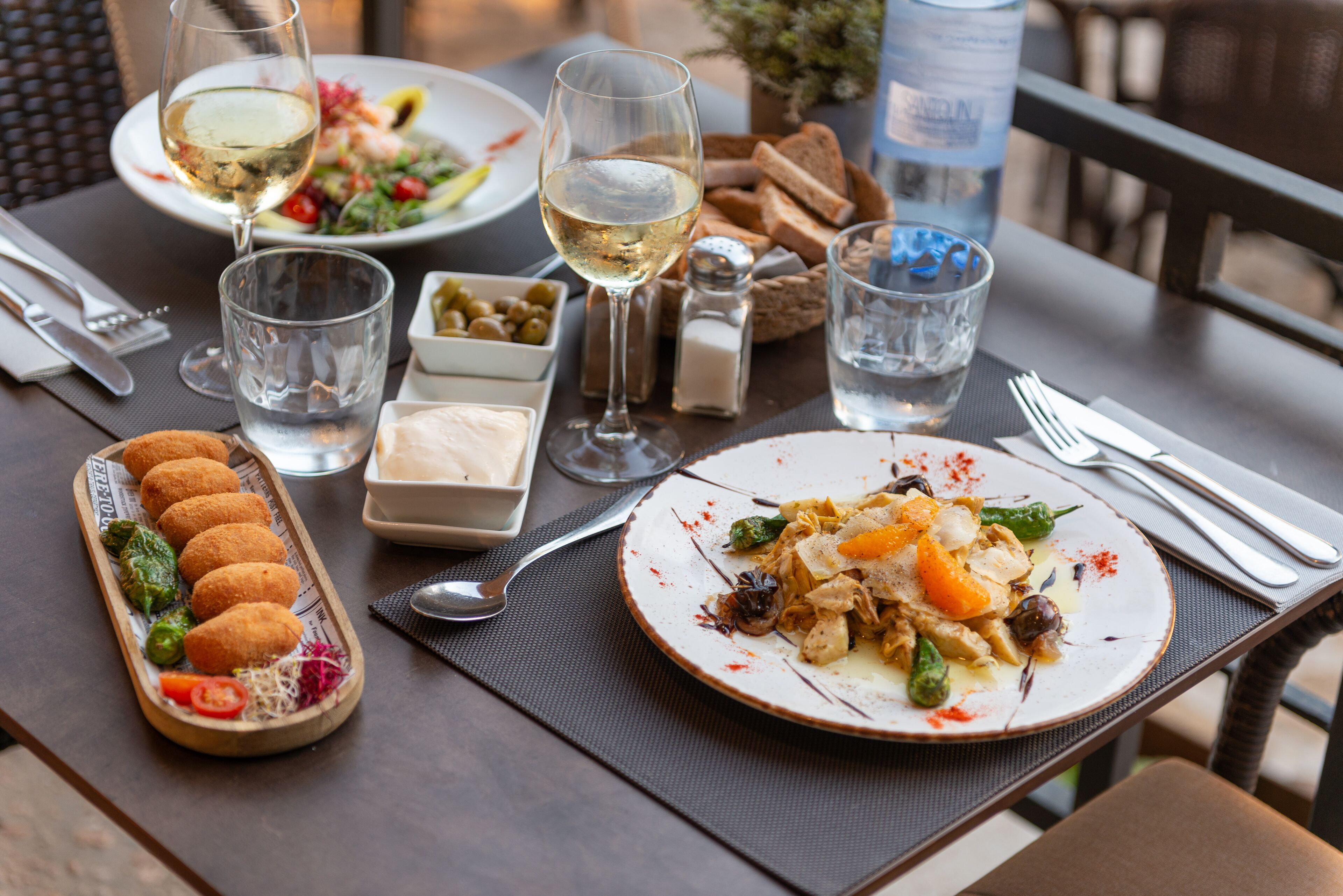 A dining table set with various dishes, including a plate of food, a glass of white wine, and a tray of bread.