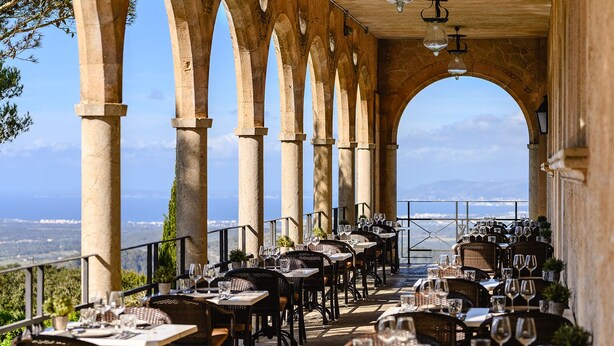 An outdoor dining area with stone arches, tables set with wine glasses, and a view of the sea.