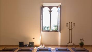 Una sala de yoga con colchonetas dispuestas en filas, un altavoz, una mesita con velas, un candelabro y una ventana con vistas a zonas verdes.