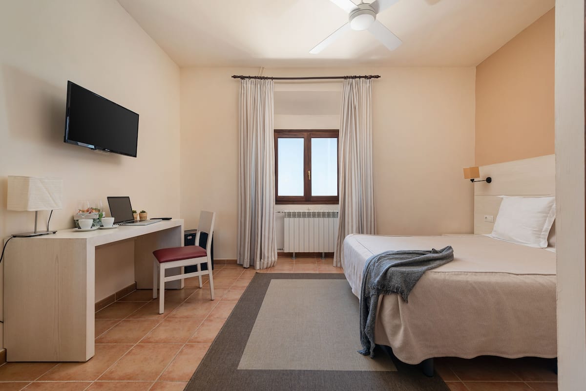 Een hotelkamer met een bed, een bureau met een laptop, een stoel, een aan de muur bevestigde televisie en een raam met gordijnen.