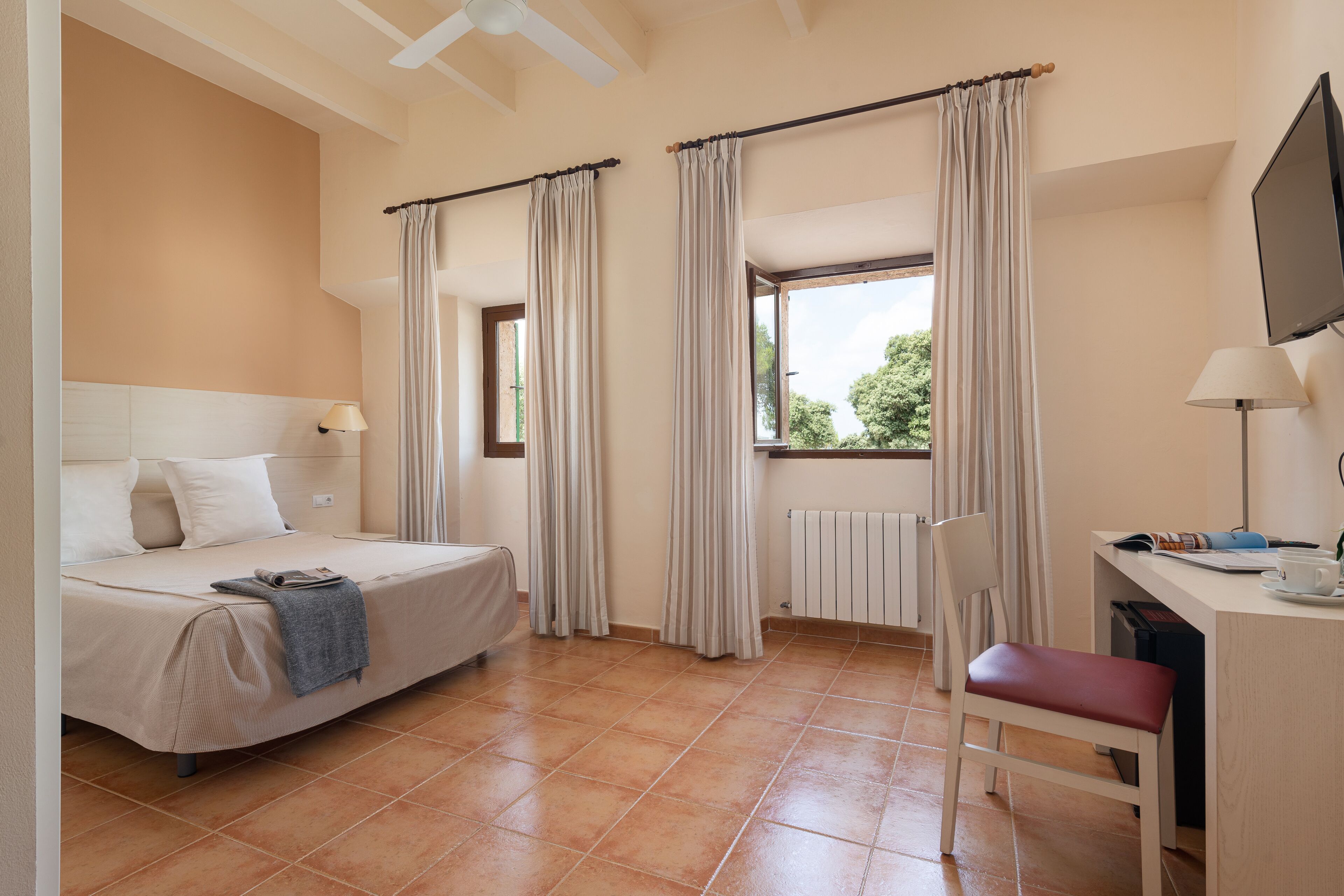 Una habitación de hotel con cama, escritorio, silla, ventana con cortinas y televisor montado en la pared.