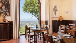 A dining area with a table set for six, a view of the countryside through an arched window, and artwork on the walls.