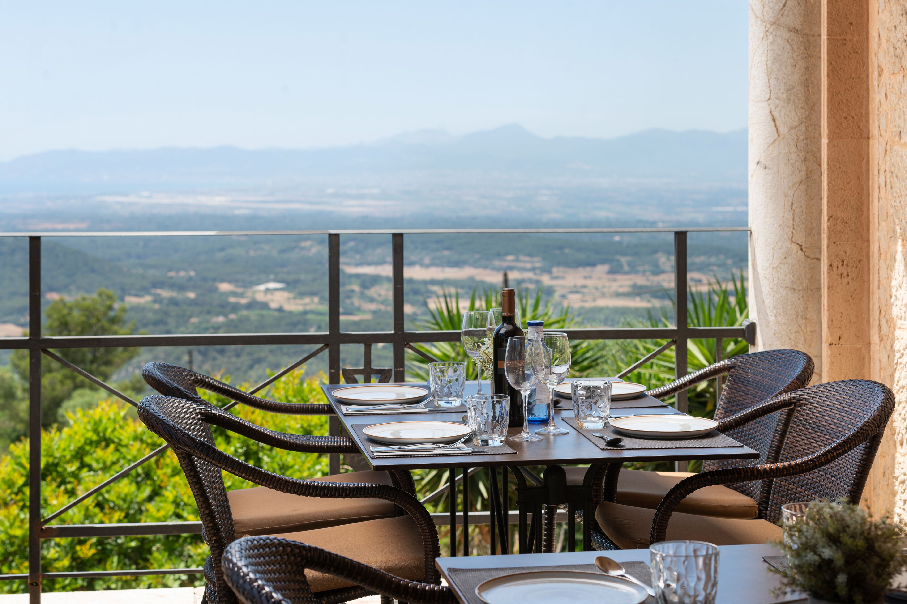 Mesa para dos personas dispuesta con sillas de mimbre, una botella de vino y una vista panorámica de montañas y vegetación.