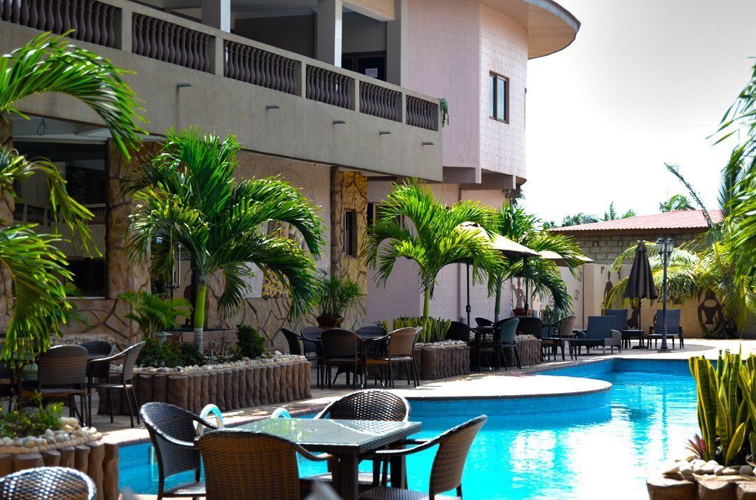 Zona de piscina con sillones y mesas, rodeada de palmeras y un edificio con balcón.