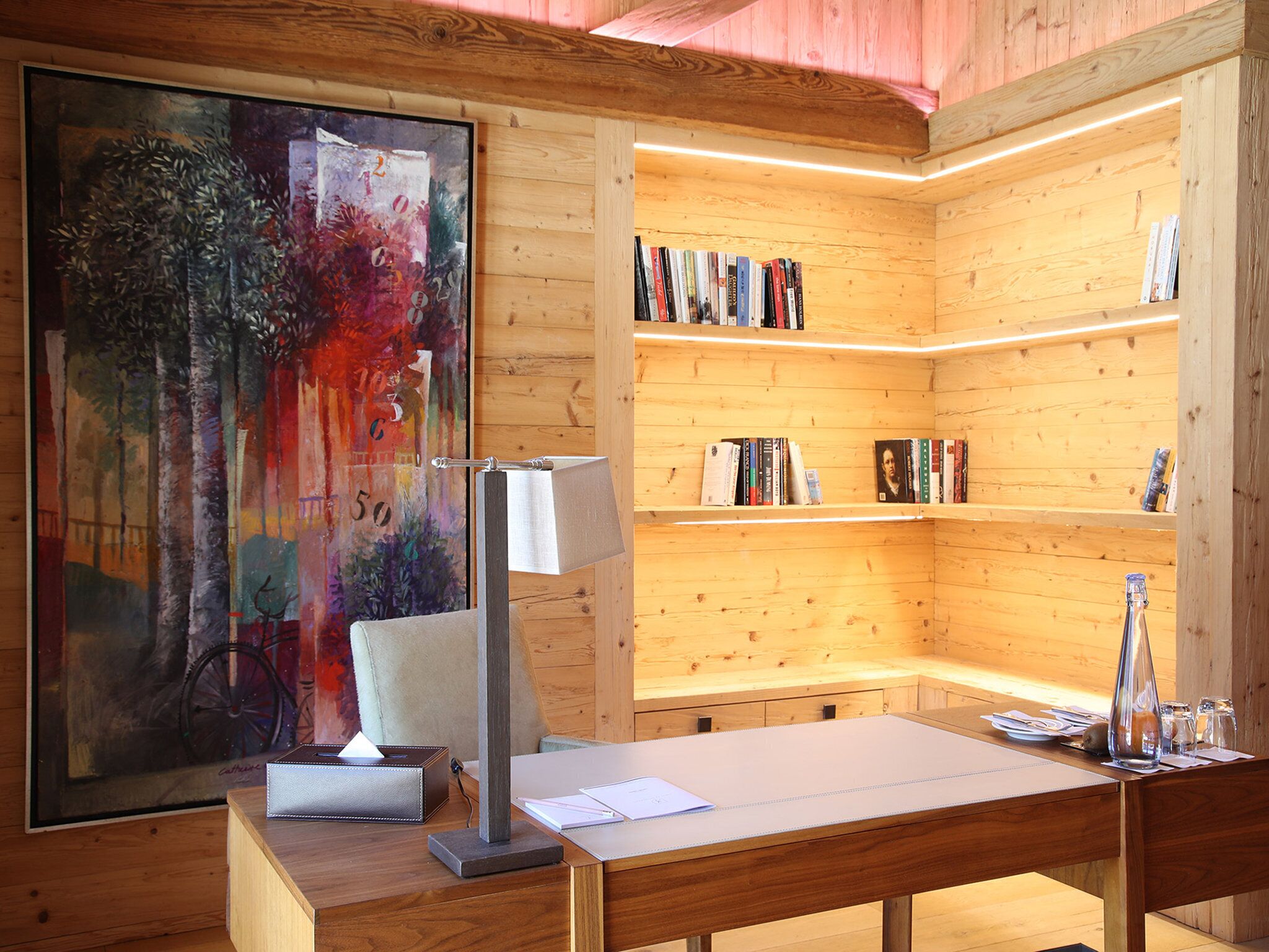 Un interior de madera con un cuadro, estanterías, un escritorio y una lámpara.