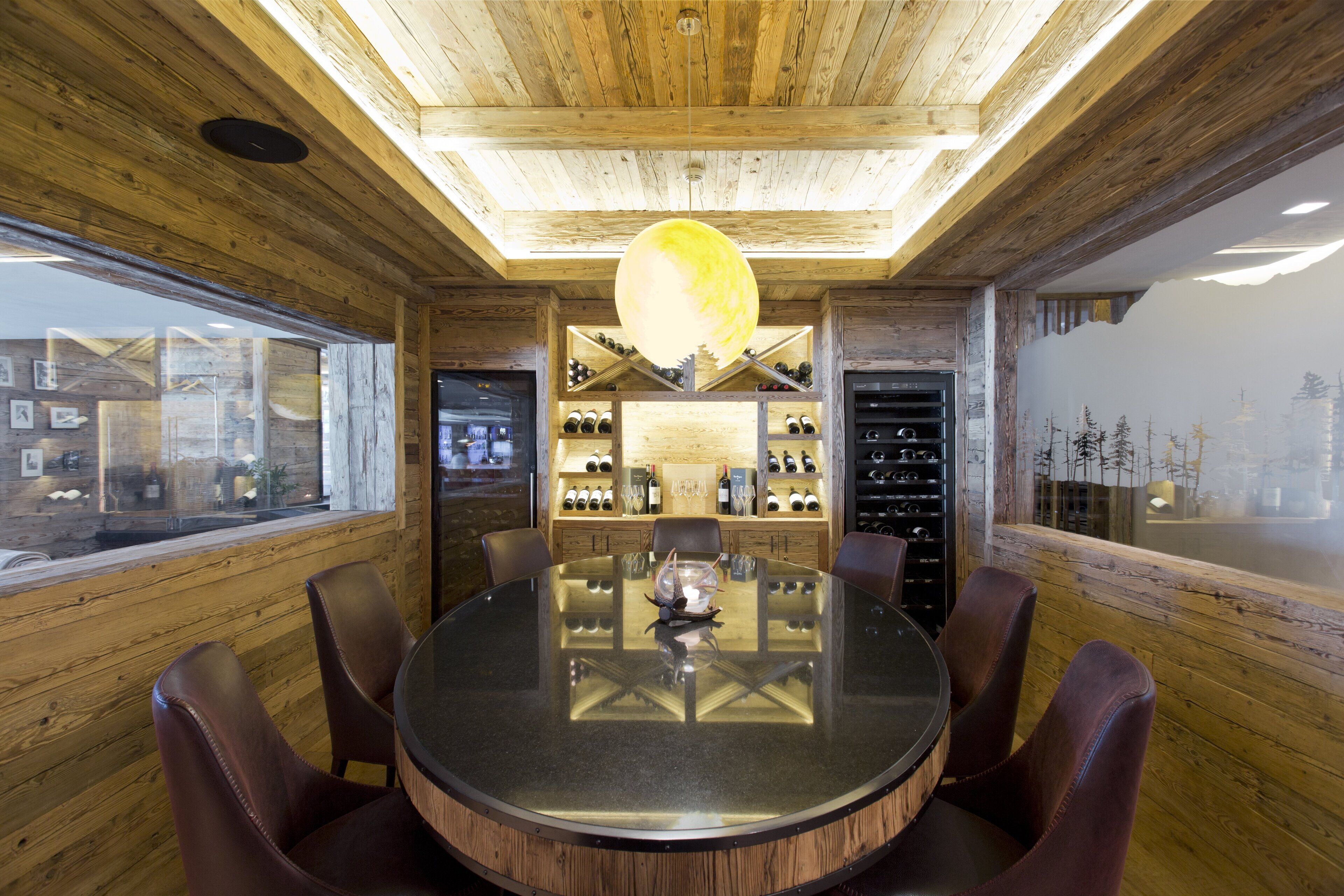 A wooden interior with a round table and chairs, a wine rack, and a large window.