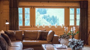 A cozy cabin interior with a wooden ceiling, a leather sofa, a coffee table with books, and a view of a white picket fence and trees through large windows. - Hotel de Rougemont & Spa (Rougemont)