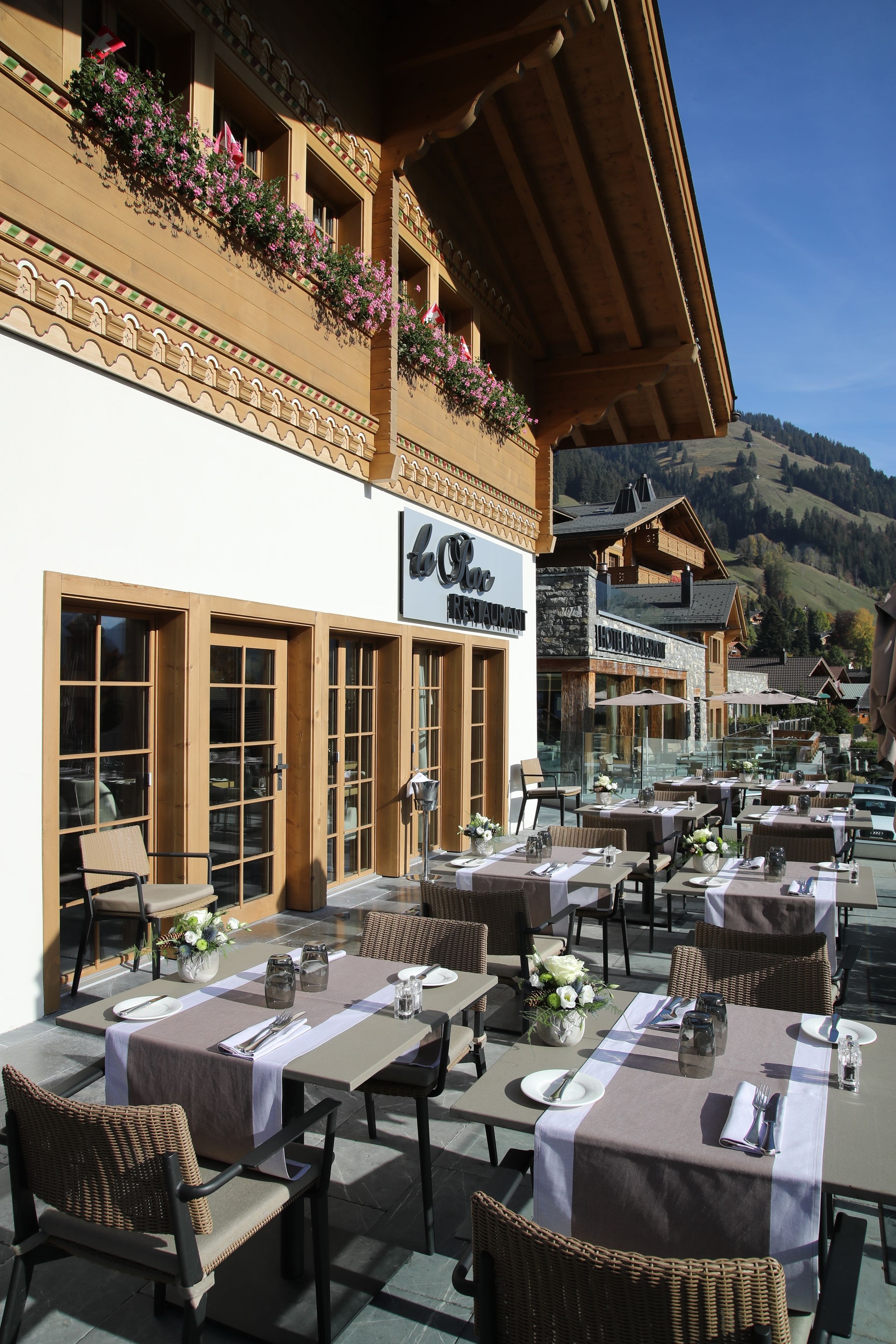 An outdoor restaurant with multiple tables and chairs, a signboard, and a mountainous backdrop.