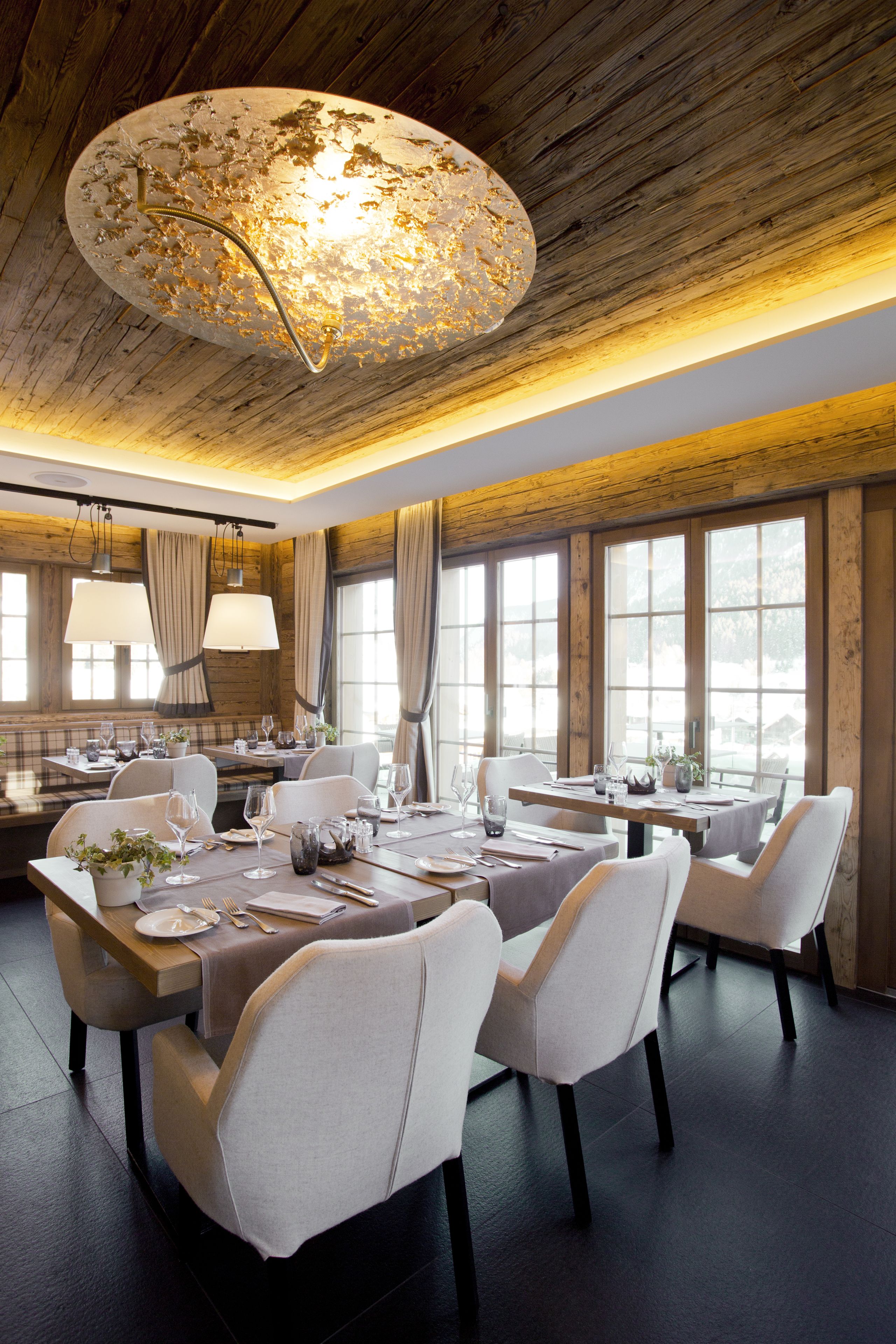 A dining area with a wooden ceiling, large windows, and a central chandelier. Tables are set with white chairs and dining ware.