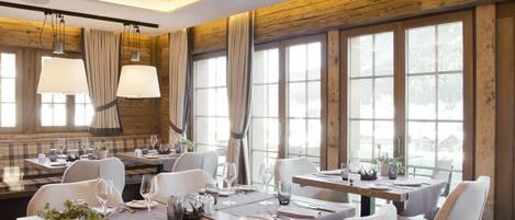 A dining area with a wooden ceiling, large windows, and a central chandelier. Tables are set with white chairs and dining ware.