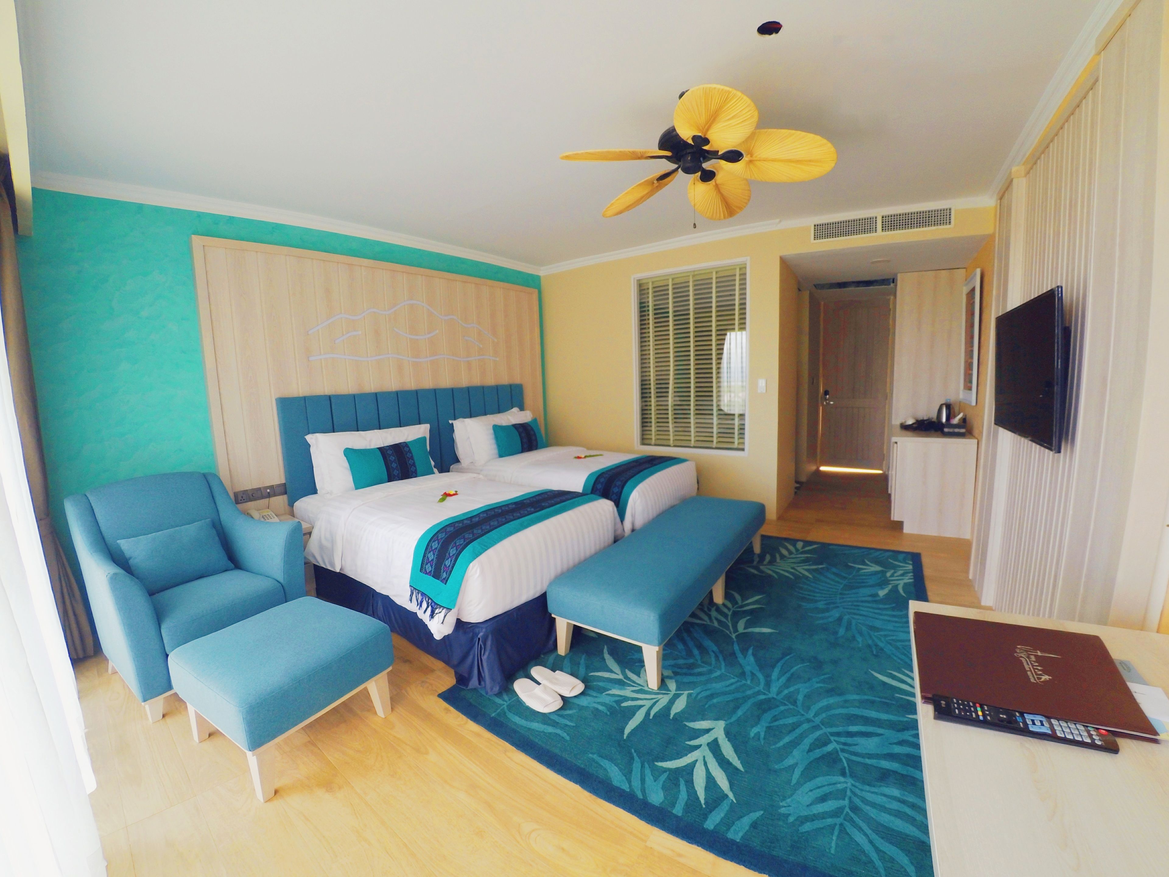 A hotel room with a bed, two armchairs, a ceiling fan, a TV, and a desk with a laptop.