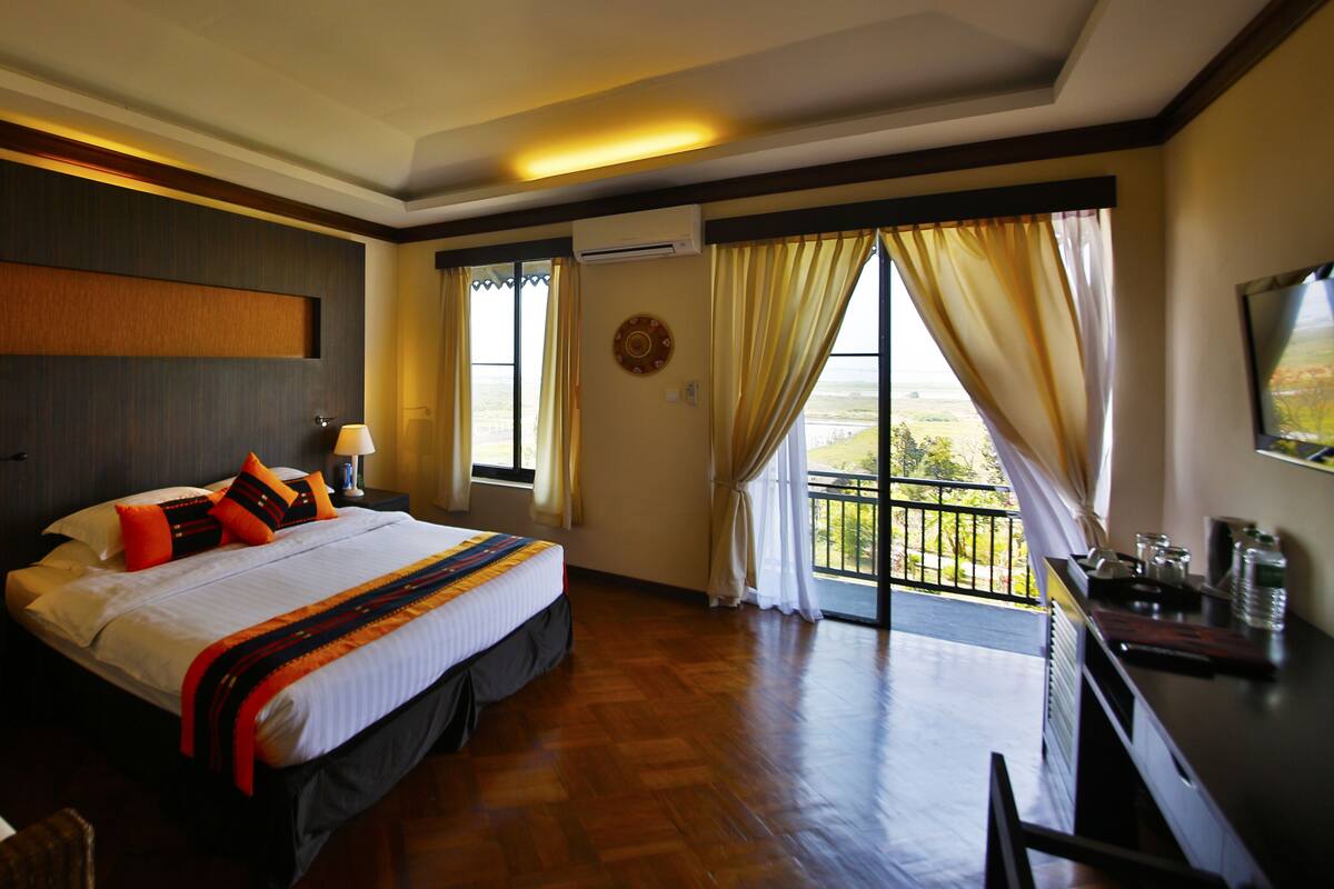 Une chambre d’hôtel avec un grand lit, un bureau avec une chaise, une fenêtre avec des rideaux et un balcon avec vue.