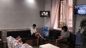 A reception area with a sofa, a small table, a TV mounted on the wall, and a window with curtains. - Legend R. G. (Rome)