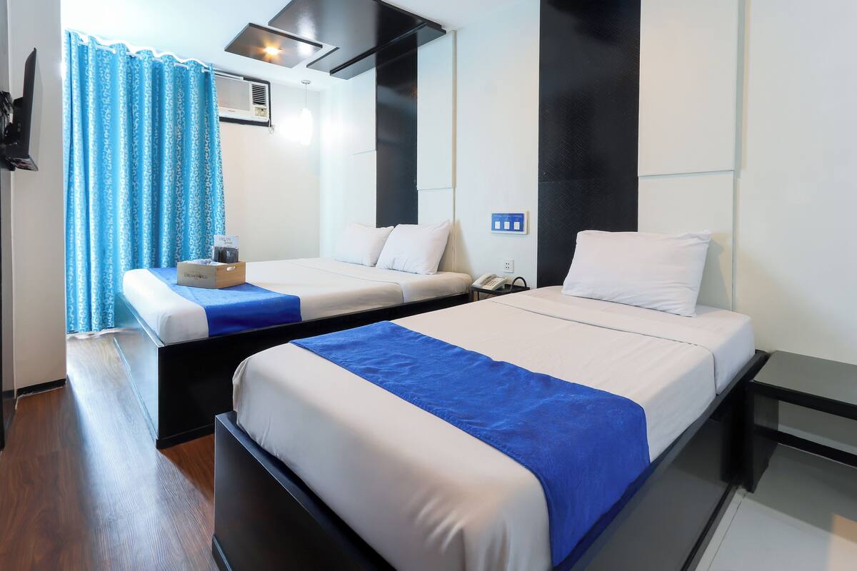 Ein Hotelzimmer mit zwei Betten, Holzboden, blauem Vorhang, einem an der Wand montierten Telefon und einem Lichtschalter.