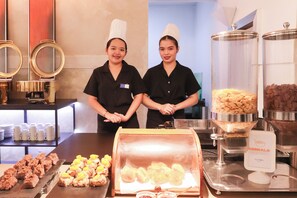 Two individuals in black uniforms standing behind a buffet counter with various food items and a sign indicating 'Cereals'. - Hotel DreamWorld North EDSA (Quezon City)