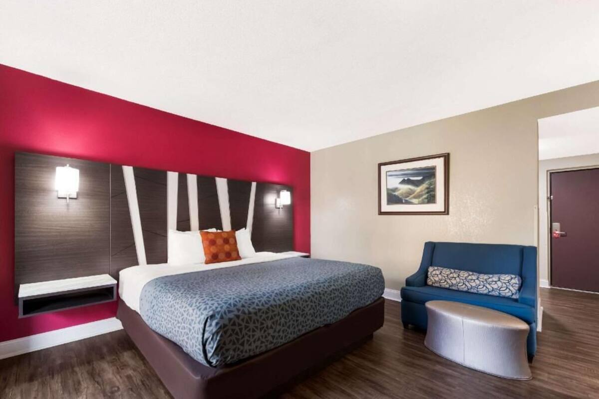 Ein Hotelzimmer mit einem Bett, einem blauen Sofa, einem kleinen Hocker, einem gerahmten Bild an der Wand und einer Tür im Hintergrund.