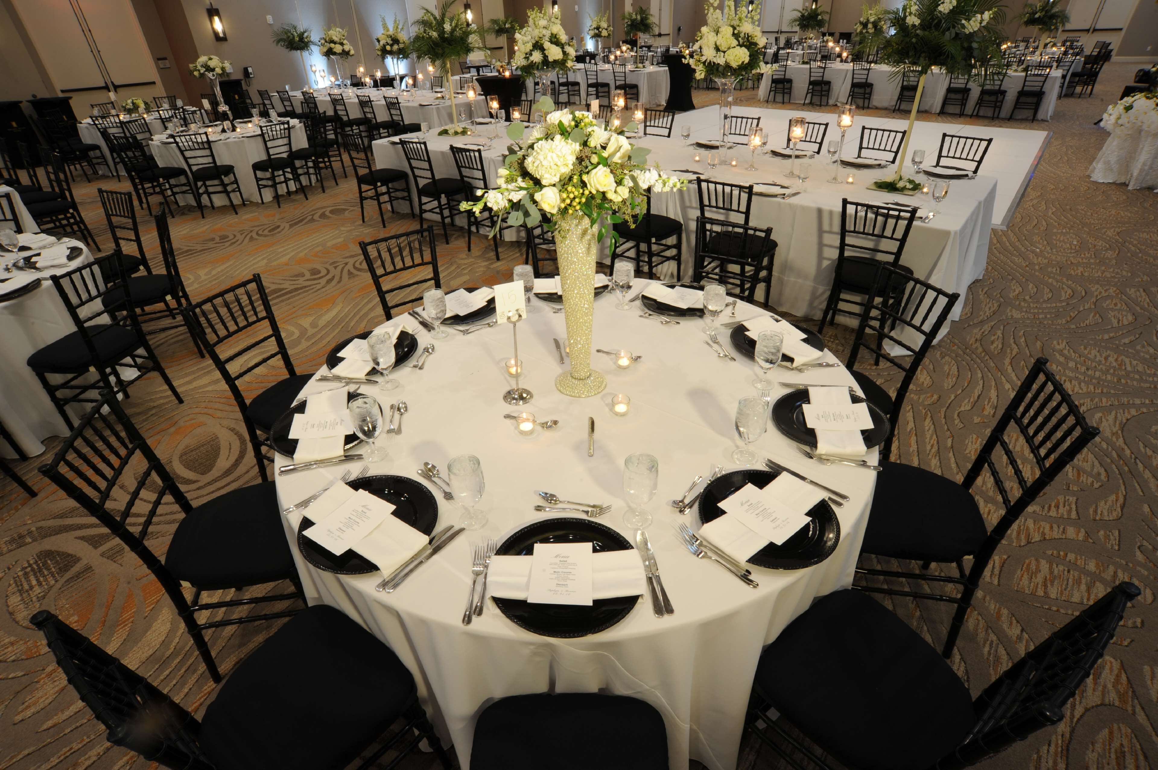 Uma sala de banquetes com mesas redondas dispostas para um evento formal, com toalhas brancas, cadeiras pretas e centros de mesa florais.