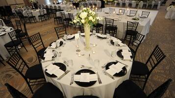 Uma sala de banquetes com mesas redondas dispostas para um evento formal, com toalhas brancas, cadeiras pretas e centros de mesa florais.