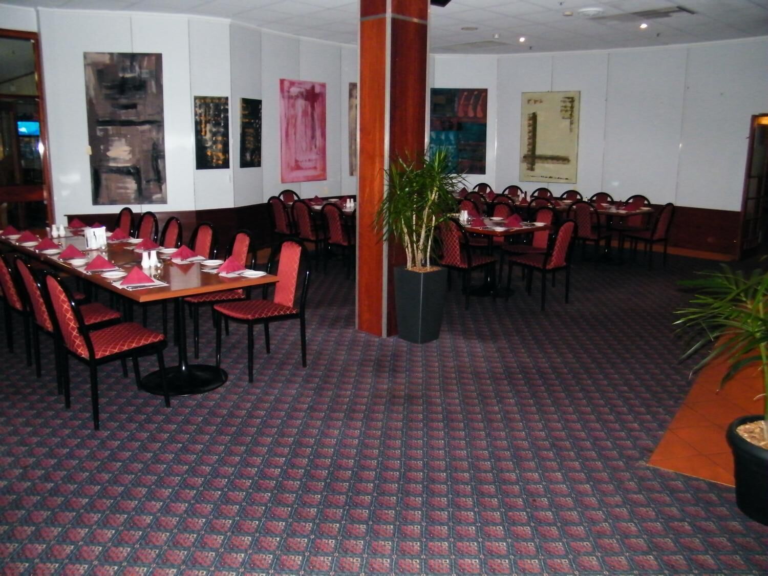 Un ristorante con sedie rosse e nere, tavoli apparecchiati con piatti bianchi e tovaglioli rossi, e un tappeto decorato.