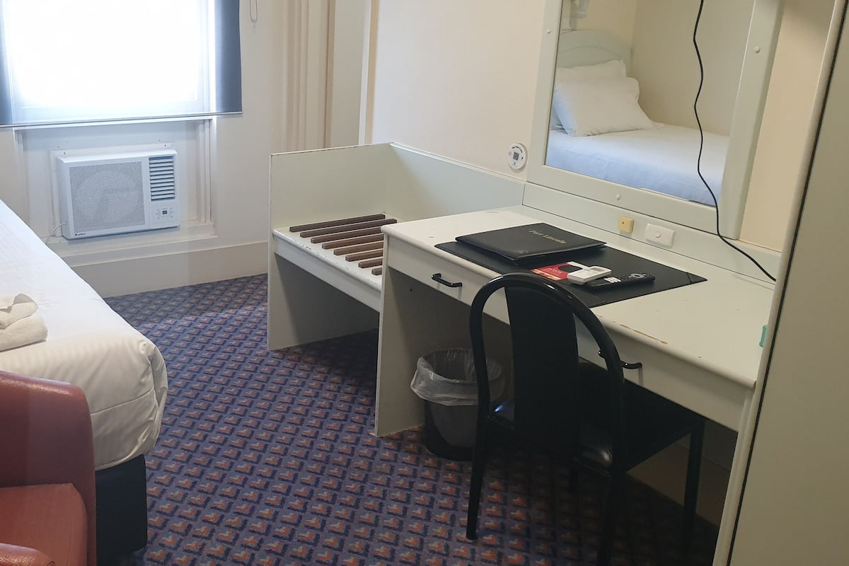 Ein Hotelzimmer mit Bett, Schreibtisch, Stuhl, Mülleimer und einem Fenster mit Klimaanlage.