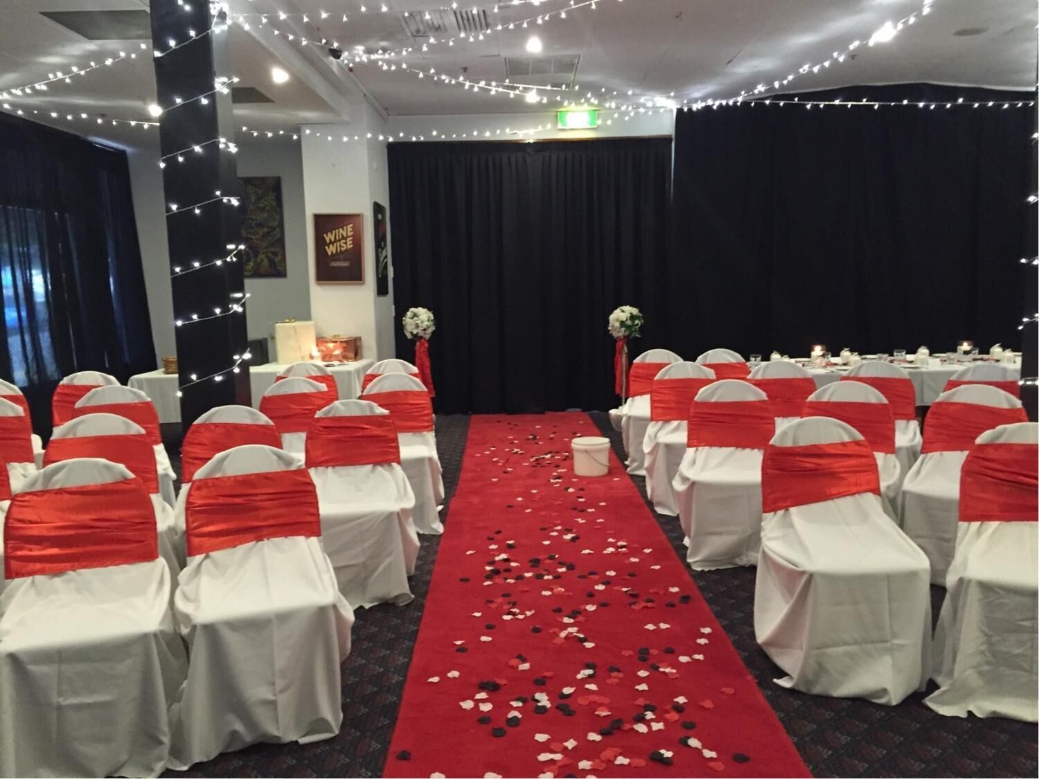 Une salle de banquet avec des rangées de tables, des nappes blanches et des rubans rouges sur les chaises.