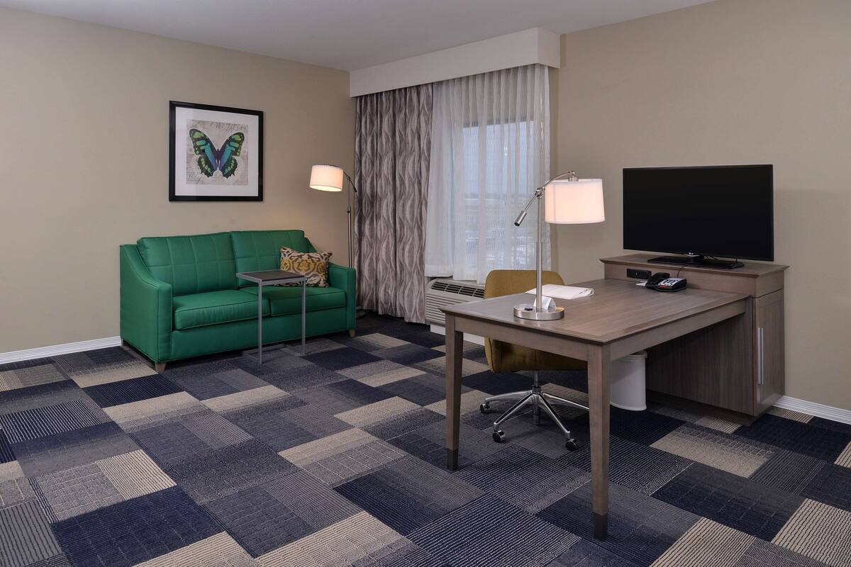 Ein Hotelzimmer mit einem grünen Sofa, einem Schreibtisch mit einem Flachbildfernseher, einer Lampe und einem gemusterten Teppich.