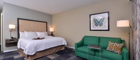 A hotel room with a bed, a green sofa, a small table, a lamp, and a framed butterfly artwork on the wall.