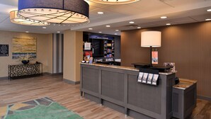 A reception area with a counter, a large lamp, a welcome sign, and a display of amenities. - Hampton Inn Broussard-Lafayette Area (Broussard)
