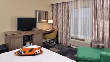 A hotel room with a bed, a desk with a television, a chair, a lamp, and an air conditioning unit.