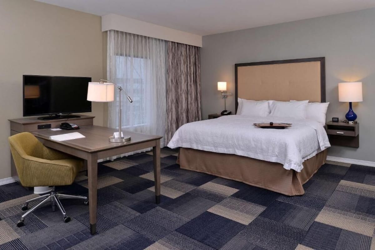 Ein Hotelzimmer mit einem Bett, einem Schreibtisch mit Fernseher, einem Sessel und zwei Nachttischlampen.