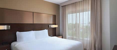 A hotel room with a large bed, a wooden headboard, a nightstand, a lamp, and a window with sheer curtains.