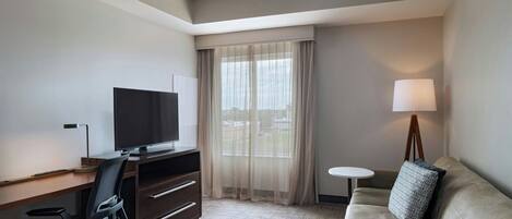 A hotel room with a desk, television, sofa, and a small table with a lamp.